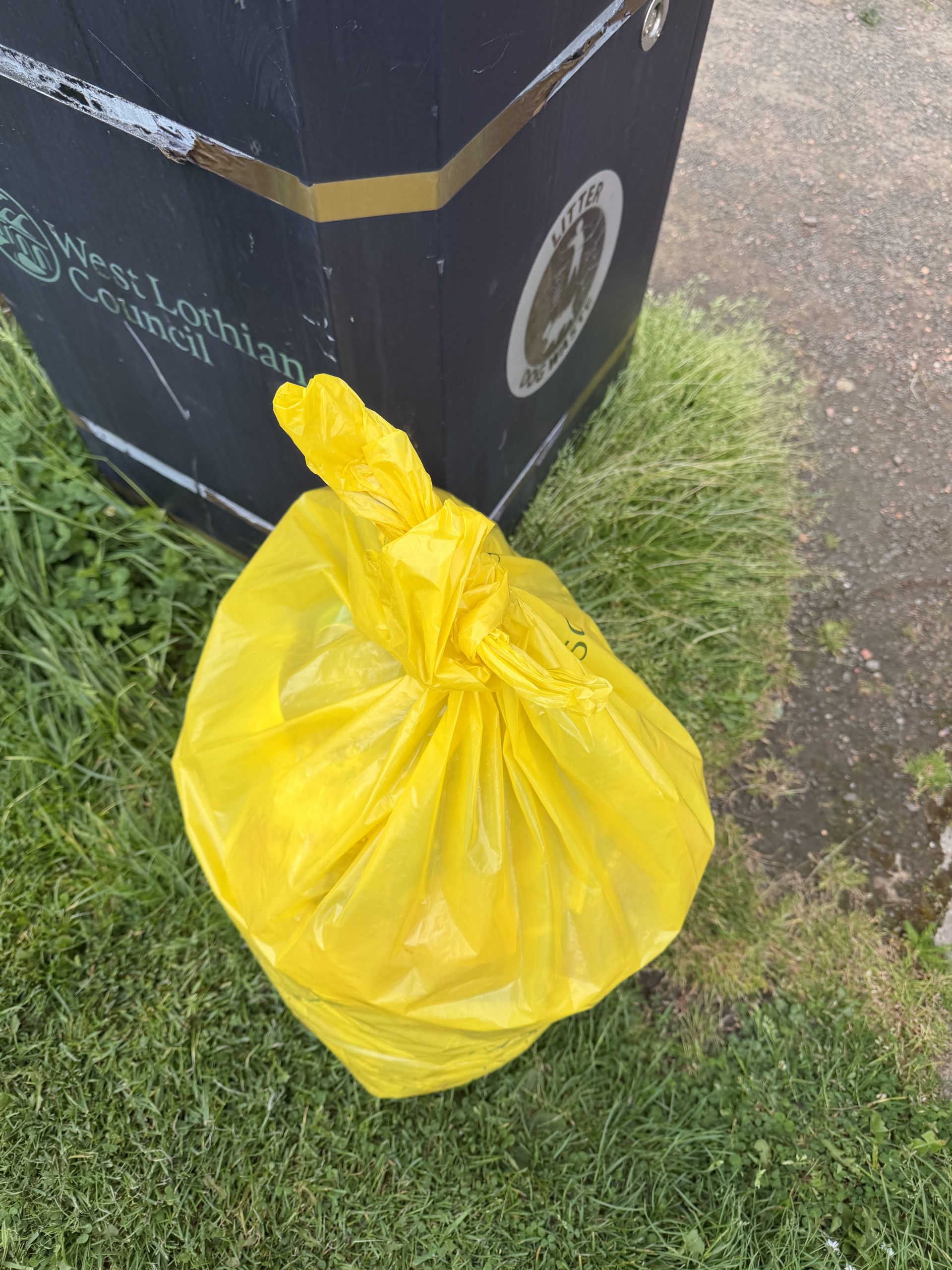 Picture showing a West Lothian Council litter bin with a full yellow litter bag beside it.